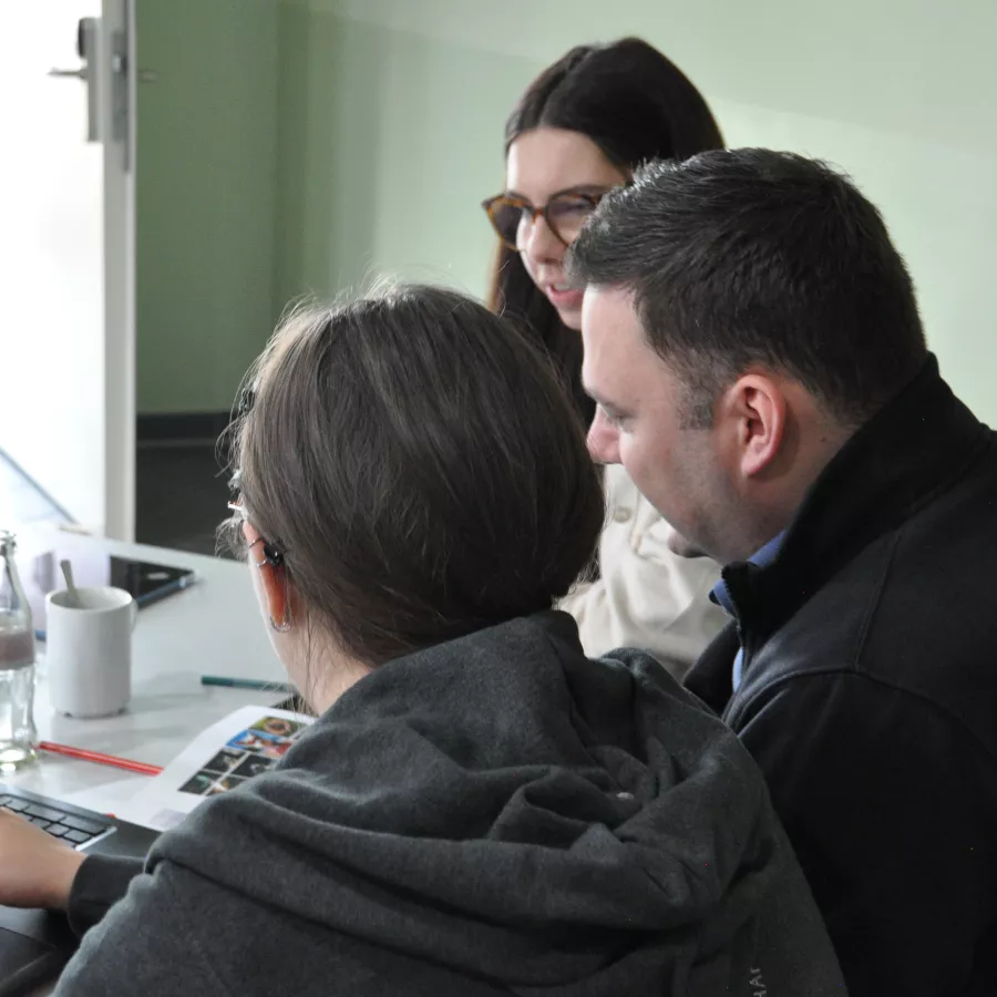 Zwei Frauen und ein Mann sitzen vor einem Laptop und arbeiten gemeinsam. 