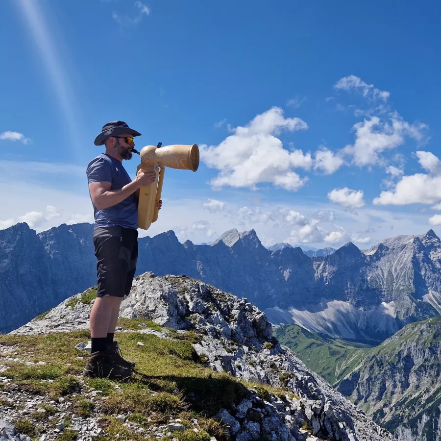 Ein Mann spielt auf einem Kompakthorn (Vogelhorn) auf einem Berg. Im Hintergrund ist ein Gebirge zu sehen. 
