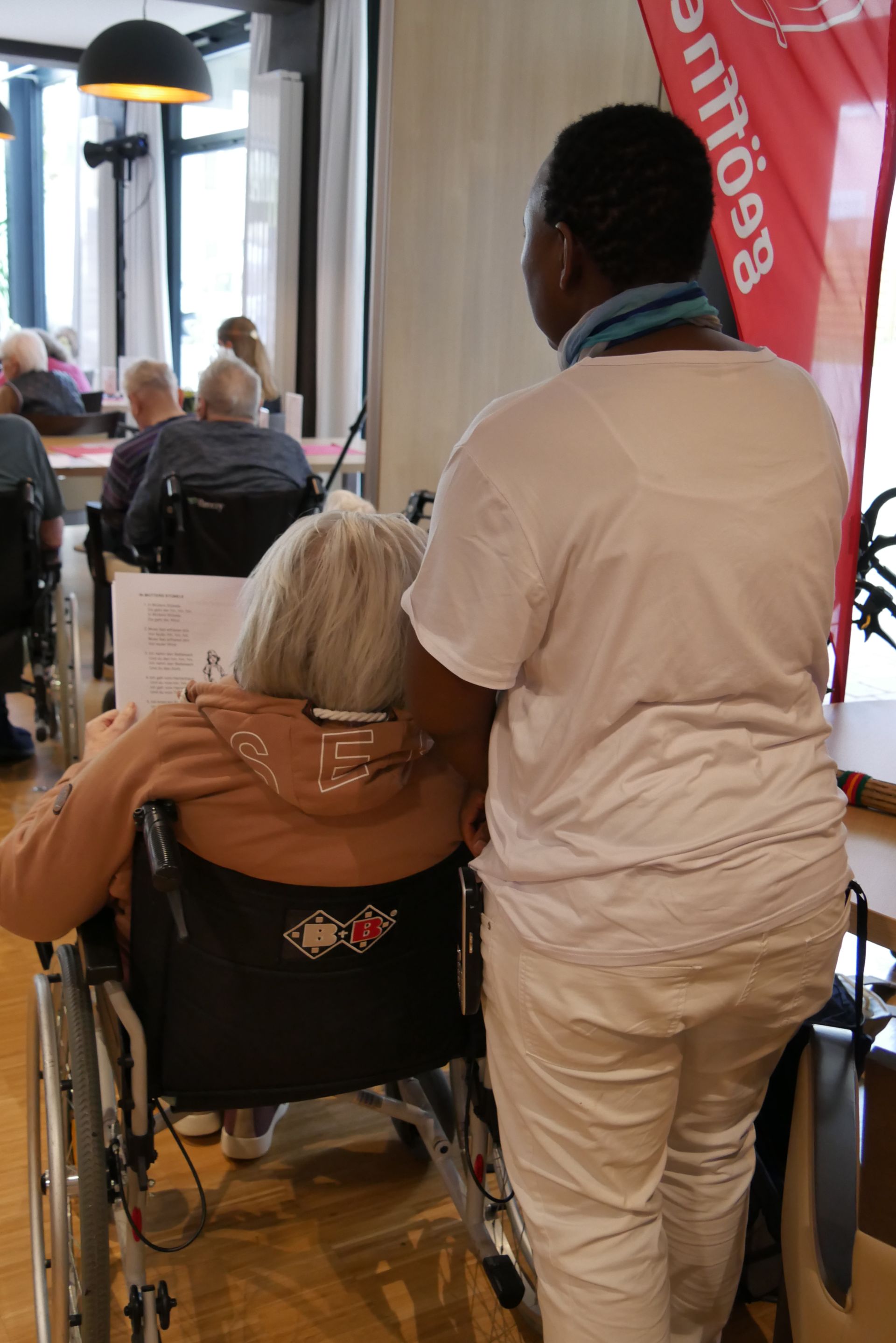 Eine Seniorin sitzt im Rollstuhl und neben ihr steht eine Pflegerin in weißer Kleidung.