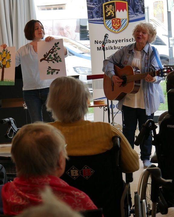 Zwei Frauen stehen nebeneinander und singen vor Publikum. eine Frau spielt dazu Gitarre. 