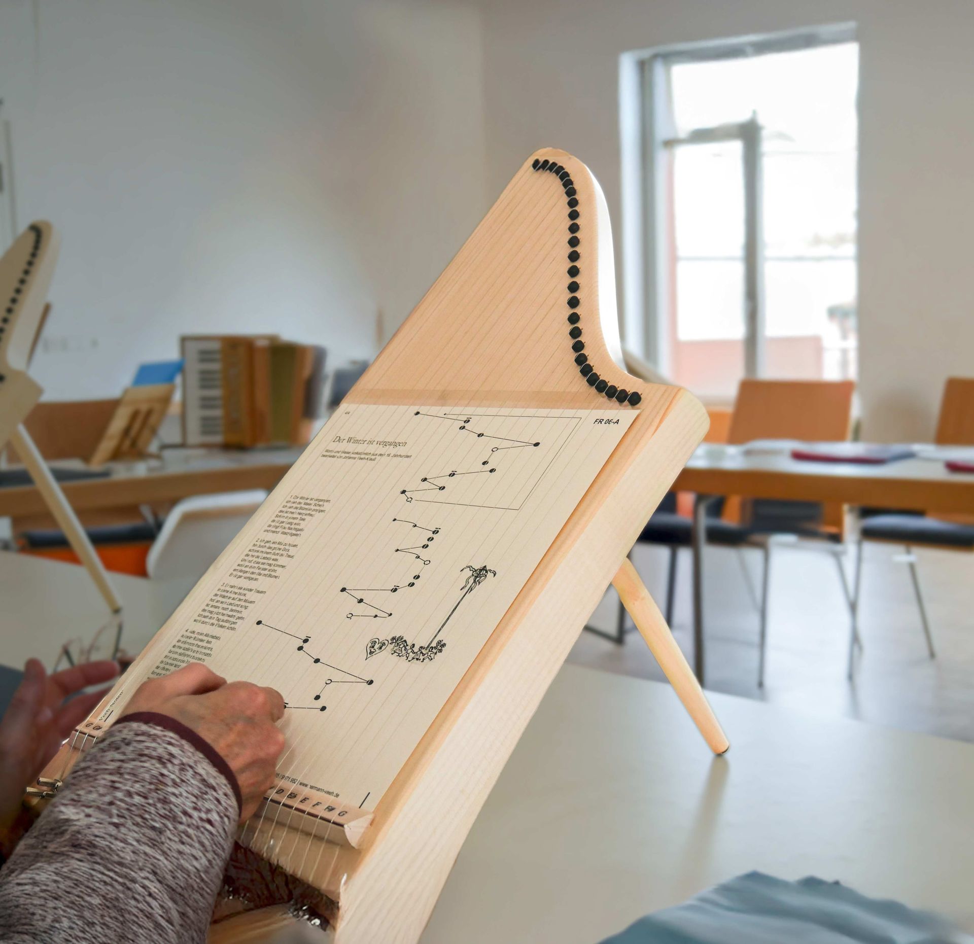 Eine Tisch∙harfe mit einem Noten∙blatt liegt auf dem Tisch. Ein Mensch zupft an einer Saite.