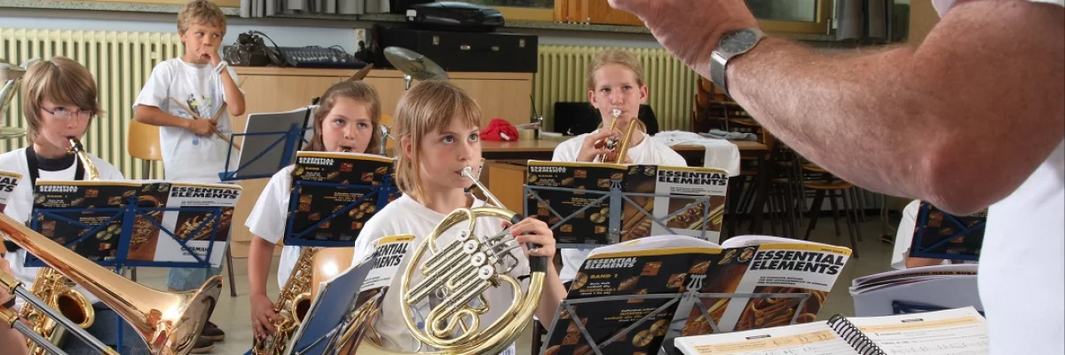 Kinder musizieren gemeinsam und werden von einem Mann dirigiert. 