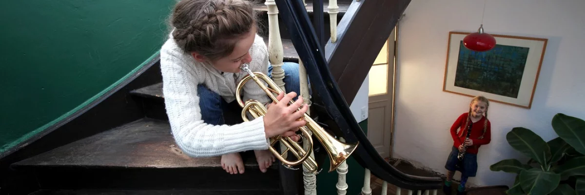 Ein Mädchen sitzt in einem Treppenhaus und spielt Trompete. Ein anderes Mädchen steht mit einer Klarinette am Fuß der Treppe.