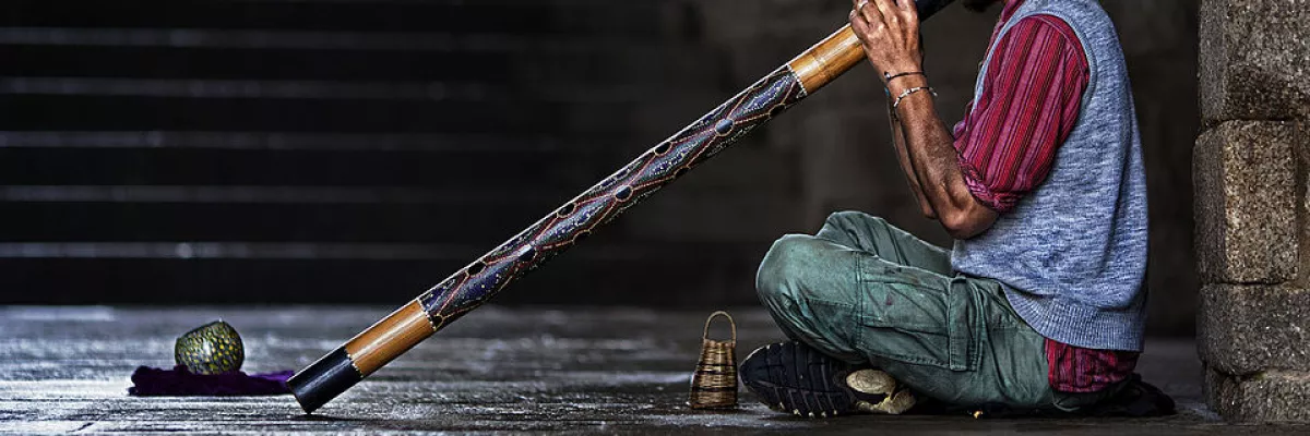 Ein Mann sitzt auf der Straße und spielt auf einem langen Didgeridoo. 