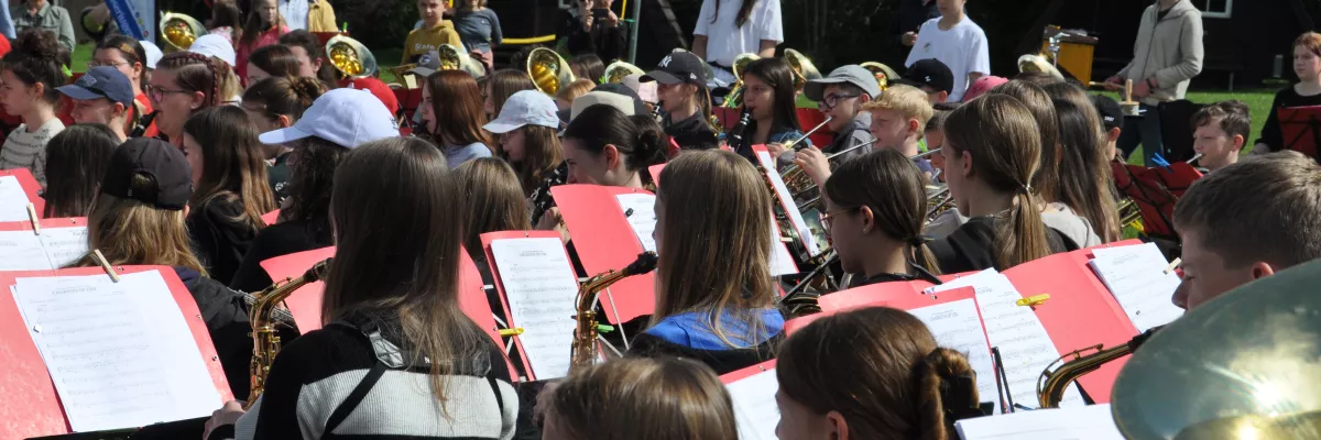Ein großes Orchester mit jungen Musiker:innen musiziert gemeinsam im Freien. Im Hintergrund sind Bäume zu sehen. 