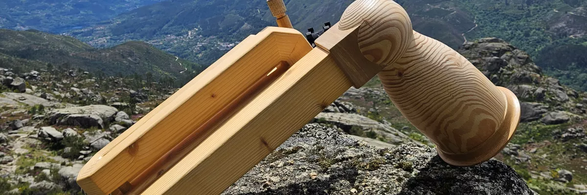 Ein Kompakthorn (Vogelhorn) aus Holz liegt auf Steinboden in der Natur. Im Hintergrund ist ein Bergpanorama zu sehen. 