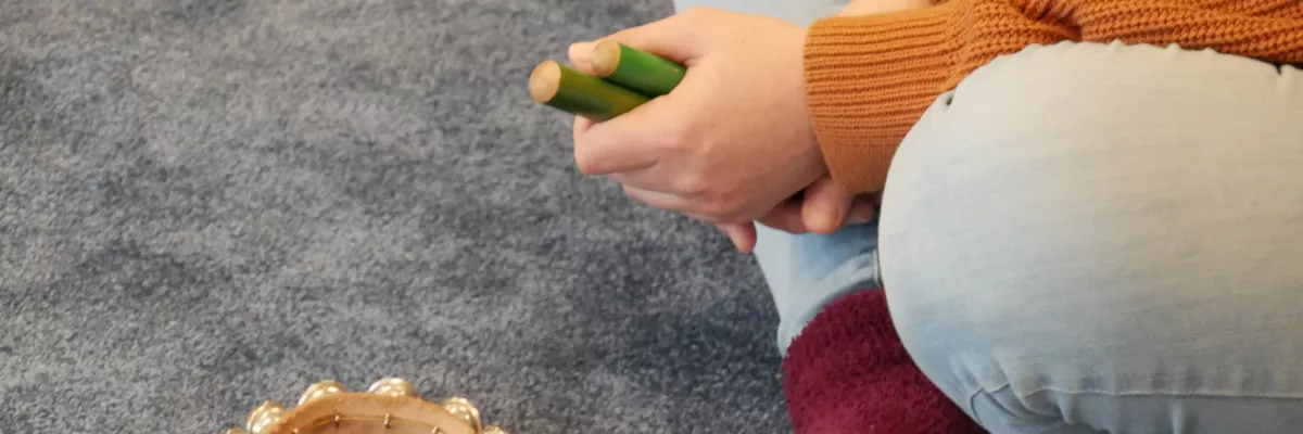 Eine Person sitzt auf einem grauen Teppich und hält Klanghölzer in der Hand. Auf dem Teppich liegt ein Schellenkranz.