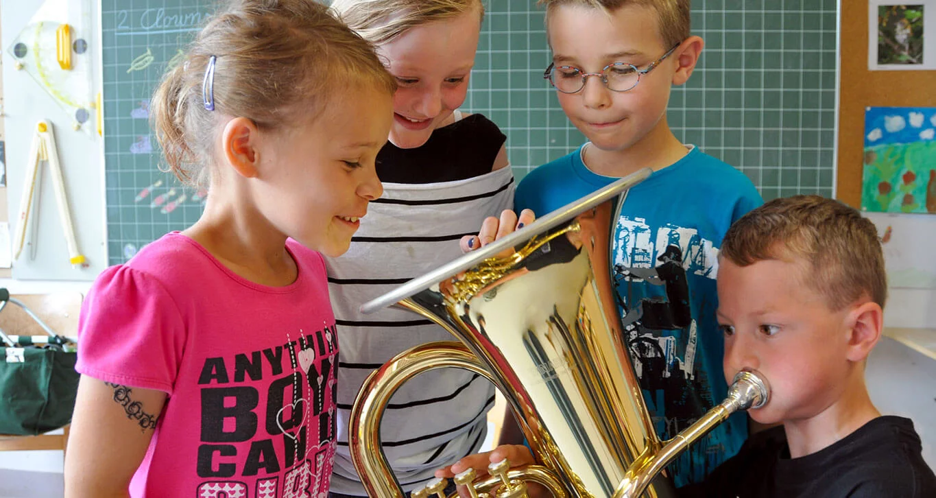 Ein Junge spielt Euphonium, drei Kindern schauen dabei in den Trichter des Euphoniums und lächeln. 