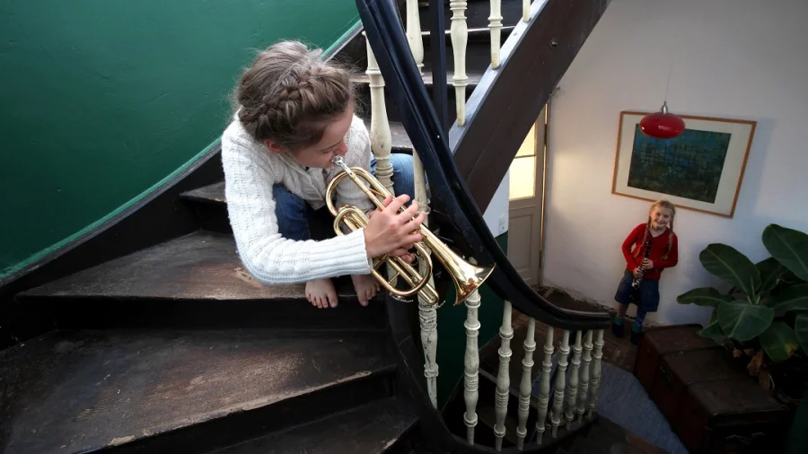Ein Mädchen sitzt in einem Treppenhaus und spielt Trompete. Ein anderes Mädchen steht mit einer Klarinette am Fuß der Treppe. 