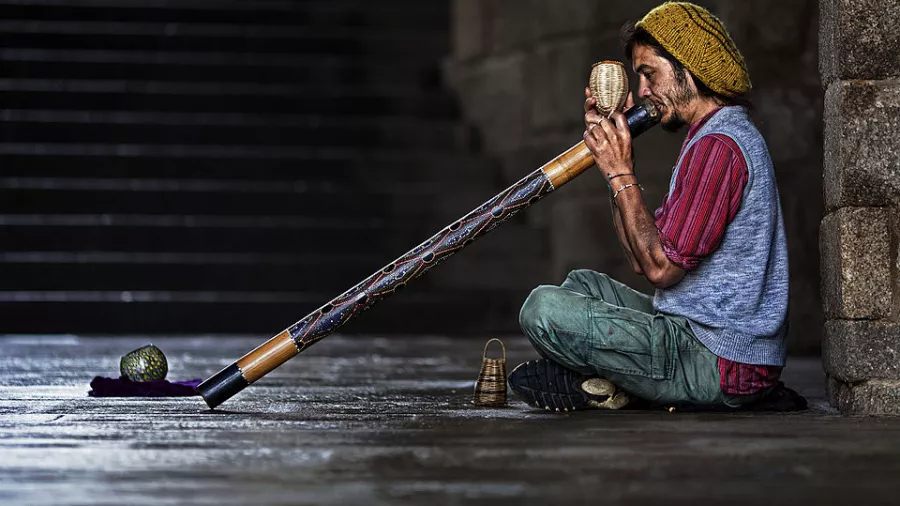 Ein Mann sitzt auf der Straße und spielt auf einem langen Didgeridoo. 