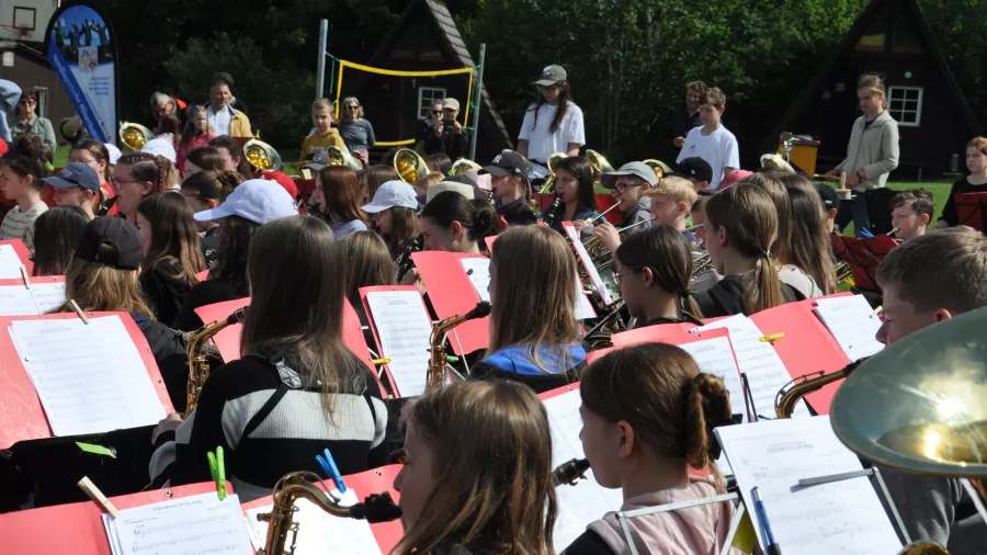 Ein großes Orchester mit jungen Musiker:innen musiziert gemeinsam im Freien. Im Hintergrund sind Bäume zu sehen.