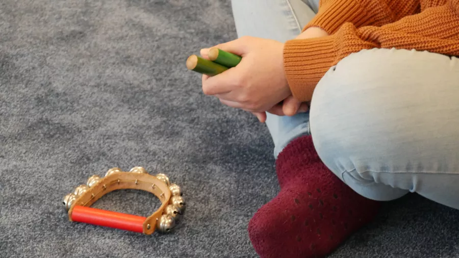 Eine Person sitzt auf einem grauen Teppich und hält Klanghölzer in der Hand. Auf dem Teppich liegt ein Schellenkranz. 