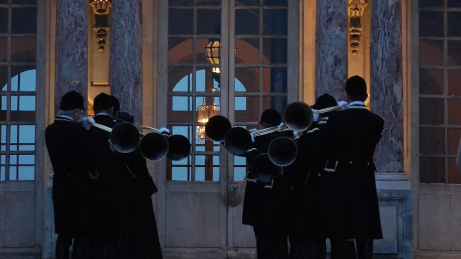 Einige Menschen in Uniform stehen in zwei Reihen vor einem Palast und spielen auf Parforcehörnern.