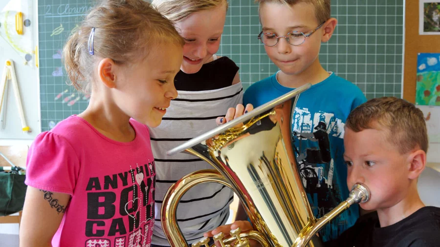 Ein Junge spielt Euphonium, drei Kindern schauen dabei in den Trichter des Euphoniums und lächeln. 