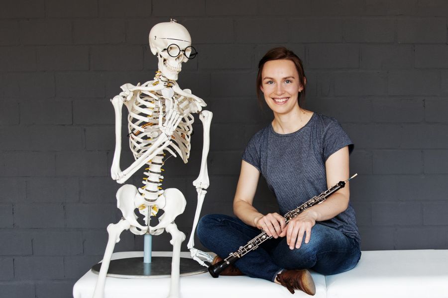 Eine Frau hält eine Oboe in der Hand und sitzt neben einem Skelett. Sie lächelt in die Kamera. 