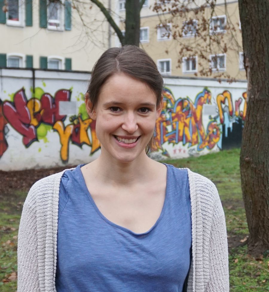 Eine Frau steht vor einer Graffiti-Wand und einer Wiese und lächelt in die Kamera. 